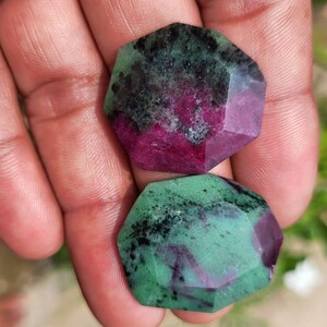 Könnte beinhalten: Zwei facettierte Edelsteine mit einer Mischung aus Farben. Der obere Stein hat eine dunkelgrüne und schwarze Oberfläche mit einem magentafarbenen Zentrum. Der untere Stein ist ähnlich, mit einer grünen und schwarzen Oberfläche und einem magentafarbenen Zentrum. Die Steine werden in einer Hand gehalten.