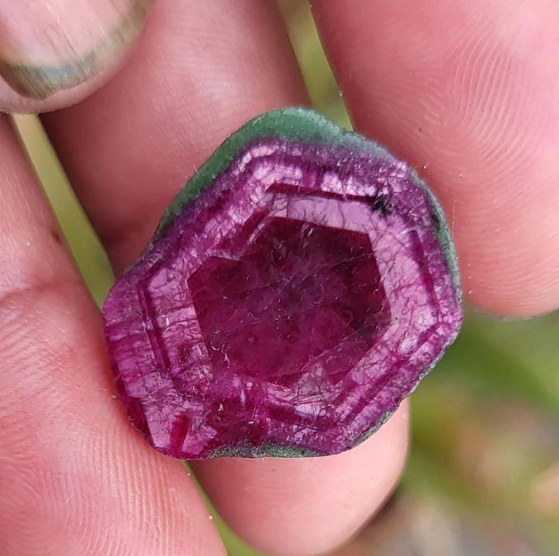 May include: A gemstone cross-section with a deep magenta core and a green outer edge. The stone features a geometric pattern of lines and facets, creating a unique texture. The stone is held in a hand, with a blurred green background.