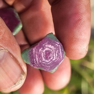 Pode incluir: Uma pedra preciosa em close-up, segurada por uma m&atilde;o. A pedra tem um centro roxo escuro com linhas brancas conc&ecirc;ntricas, rodeado por uma borda verde. O padr&atilde;o &uacute;nico da pedra &eacute; vis&iacute;vel.