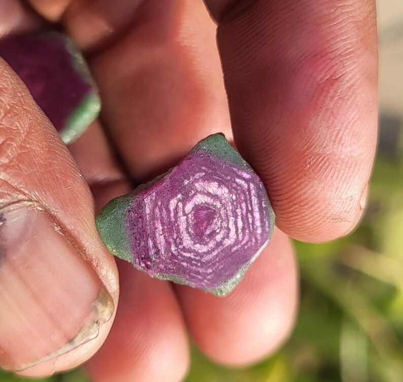 Pode incluir: Uma pedra preciosa em close-up, segurada por uma m&atilde;o. A pedra tem um centro roxo escuro com linhas brancas conc&ecirc;ntricas, rodeado por uma borda verde. O padr&atilde;o &uacute;nico da pedra &eacute; vis&iacute;vel.