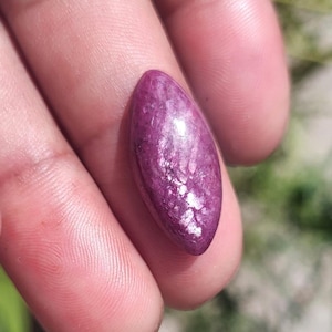 Op de afbeelding: Een gepolijste, ovaalvormige edelsteen in tinten diep paars en magenta. De steen heeft een textuur, kristallijn uiterlijk met lichtere gebieden, waardoor een glinsterend effect ontstaat. De edelsteen wordt in een hand gehouden.