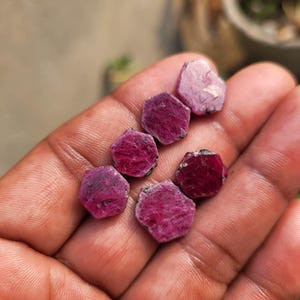 May include: A collection of raw, uncut ruby gemstones. The gemstones are a deep red-purple color, with a rough, natural texture. The rubies are in various shapes and sizes, and are displayed in a hand.