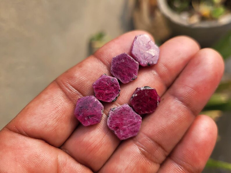 May include: A collection of raw, uncut ruby gemstones. The gemstones are a deep red-purple color, with a rough, natural texture. The rubies are in various shapes and sizes, and are displayed in a hand.