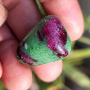 May include: A polished gemstone with a mix of colors. The stone has a vibrant green base with patches of deep red and purple. The smooth, rounded shape of the stone is visible.