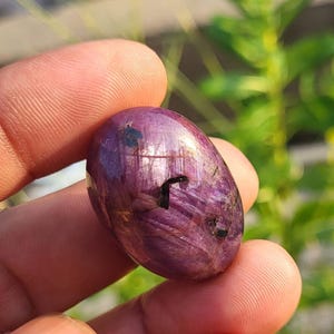 Könnte beinhalten: Ein polierter, oval geformter Rubin-Edelstein in einem tiefen Magenta- und Violettton. Der Stein zeigt eine glatte, reflektierende Oberfläche mit sichtbaren internen Kristallstrukturen, natürlichen Einschlüssen und einem markanten dunklen, kantigen Riss.