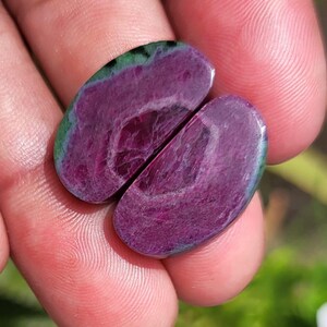 K&ouml;nnte beinhalten: Zwei ovale Edelsteine ​​in tiefem Magenta mit einem Wirbelmuster. Die Steine ​​haben einen gr&uuml;nen und blauen Rand. Die Edelsteine ​​werden in einer Hand gehalten, wobei der Hintergrund unscharf ist.