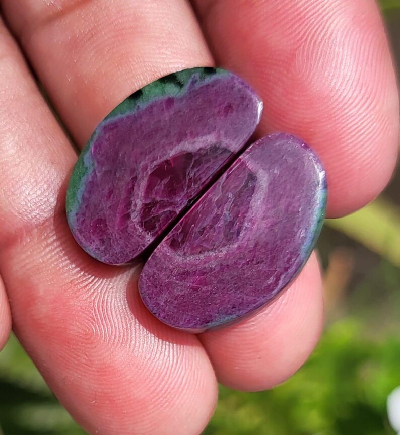 K&ouml;nnte beinhalten: Zwei ovale Edelsteine ​​in tiefem Magenta mit einem Wirbelmuster. Die Steine ​​haben einen gr&uuml;nen und blauen Rand. Die Edelsteine ​​werden in einer Hand gehalten, wobei der Hintergrund unscharf ist.