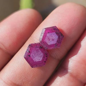 May include: Two hexagonal, faceted gemstones with a deep magenta hue are displayed. The gems have a lighter center and darker edges with small black inclusions. The gems are held in a hand, showcasing their polished surfaces and geometric shape.
