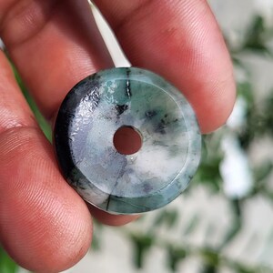 May include: A circular, donut-shaped pendant made of polished green and white stone with a smooth, glossy finish. The piece features a central hole and displays natural color variations, including dark green, light green, and white marbled patterns.