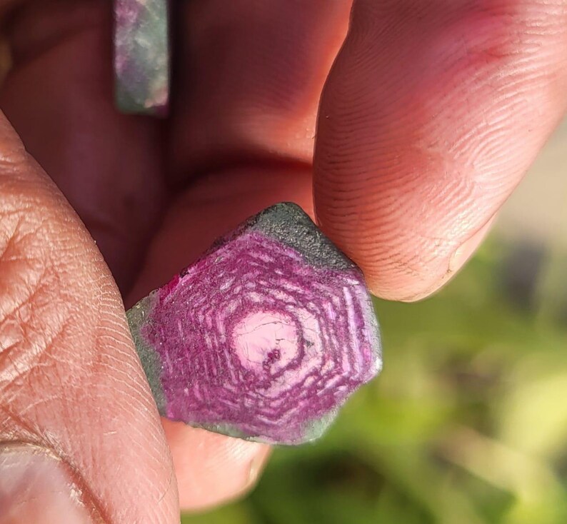 Pode incluir: Close-up de uma pedra preciosa segurada entre os dedos. A pedra tem um interior rosa e roxo com um padr&atilde;o geom&eacute;trico, rodeado por uma borda verde escura e preta. A superf&iacute;cie da pedra &eacute; &aacute;spera e texturizada.