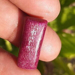 May include: A rectangular, polished gemstone in a deep magenta hue. The stone has a textured surface with visible graining. The gemstone is held between two fingers, showcasing its size and color.