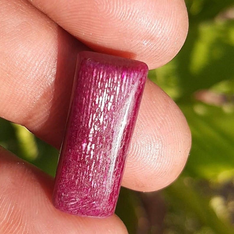 May include: A rectangular, polished gemstone in a deep magenta hue. The stone has a textured surface with visible graining. The gemstone is held between two fingers, showcasing its size and color.