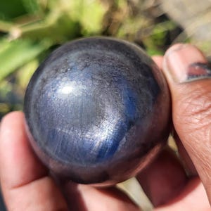 May include: A dark blue sphere with a polished, reflective surface. The sphere is held in a hand, with the background blurred. The sphere's color shifts with light, showing hints of silver and blue. A decorative item.