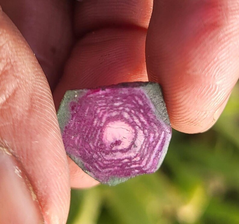 Pode incluir: Um close-up de uma pedra preciosa com um padr&atilde;o marcante. A pedra apresenta c&iacute;rculos conc&ecirc;ntricos de rosa intenso e branco, com uma borda verde. A pedra preciosa &eacute; segurada entre os dedos, mostrando sua estrutura interna &uacute;nica e varia&ccedil;&otilde;es de cor.