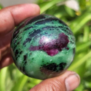 May include: A polished gemstone sphere with a mix of colours. The sphere has a base of green with black and dark red inclusions. The sphere is held in a hand against a blurred green background.