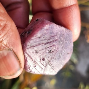May include: A rough-cut, faceted ruby gemstone with a deep pinkish-red hue. The surface displays natural lines and imperfections, held between fingers. The gemstone is a natural mineral, suitable for jewelry making or collecting.
