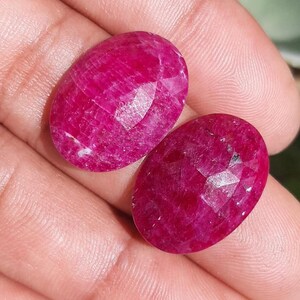 May include: Two oval-shaped, faceted rubies. The gemstones are a deep, vibrant red color. The facets catch the light, creating a sparkling effect. The rubies are displayed against a neutral background.