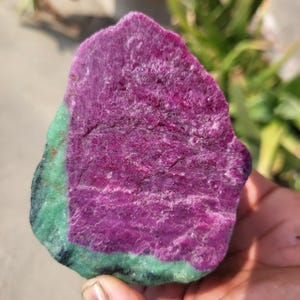 May include: A natural gemstone with a striking magenta core and a green edge. The stone's surface is textured and irregular, highlighting its raw, unrefined form. The colours are vivid and intense, showcasing the natural beauty of the mineral.