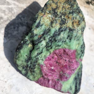 May include: A rough-cut gemstone featuring a vibrant mix of colors. The stone displays a base of green and black, with a prominent patch of deep pink-red. The natural rock has a textured surface and irregular shape.