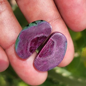 K&ouml;nnte beinhalten: Zwei polierte, oval geformte Edelsteine in tiefem Magenta mit einer geschichteten Innenstruktur. Ein Stein hat einen gr&uuml;nen und blauen Rand. Die Edelsteine werden in einer Hand gehalten und zeigen ihre nat&uuml;rliche Sch&ouml;nheit.