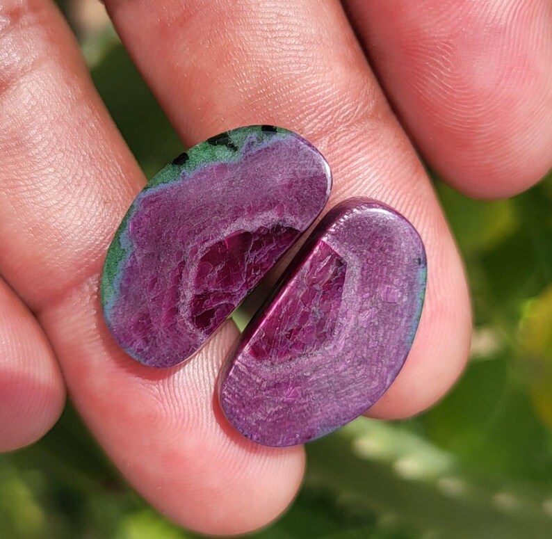 K&ouml;nnte beinhalten: Zwei polierte, oval geformte Edelsteine in tiefem Magenta mit einer geschichteten Innenstruktur. Ein Stein hat einen gr&uuml;nen und blauen Rand. Die Edelsteine werden in einer Hand gehalten und zeigen ihre nat&uuml;rliche Sch&ouml;nheit.
