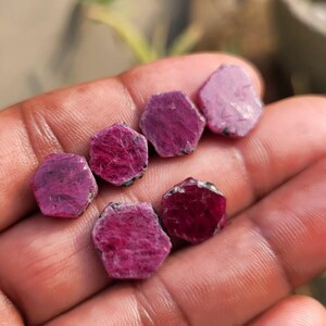 May include: A collection of raw ruby gemstones, each with a hexagonal shape and varying shades of deep red and pink. The rough, natural texture of the stones is visible, with some showing dark inclusions. The gemstones are held in a hand, showcasing their natural beauty.