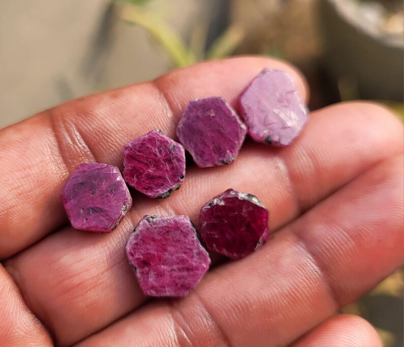 May include: A collection of raw ruby gemstones, each with a hexagonal shape and varying shades of deep red and pink. The rough, natural texture of the stones is visible, with some showing dark inclusions. The gemstones are held in a hand, showcasing their natural beauty.