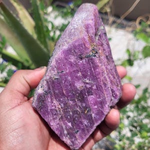 May include: A large, raw, purple crystal with a rough, textured surface. The crystal is held in a hand, showcasing its natural, unpolished form. The color is a deep, vibrant purple with some darker inclusions.