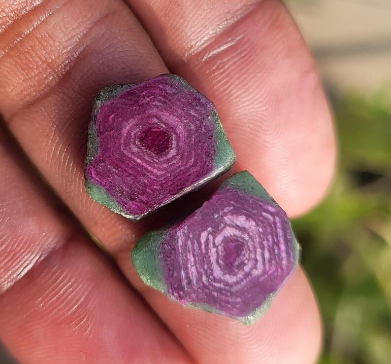 Pode incluir: Duas pedras preciosas em bruto com um centro magenta profundo e uma borda verde. As pedras preciosas t&ecirc;m um padr&atilde;o geom&eacute;trico e s&atilde;o seguradas em uma m&atilde;o, com um fundo verde desfocado.