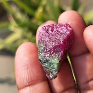 以下が含まれることがあります： 鮮やかな色のグラデーションを持つ、未加工の宝石。上部は濃いマゼンタ色で、下部に向かって緑色に変化しています。石は質感があり、自然な外観をしています。