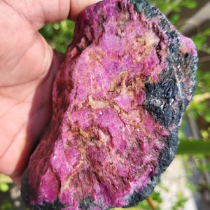 Puede incluir: Una gema de rubí en bruto, sin cortar, de color rosa vibrante, acentuada por zonas negras y verdes. La piedra natural tiene una superficie texturizada, mostrando su estado bruto y sin pulir. Una pieza única para coleccionistas.