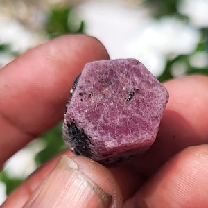 Pode incluir: Um espécime de cristal bruto de forma hexagonal com uma superfície superior em rosa magenta profundo e uma estrutura cristalina texturizada. Os lados do mineral exibem inclusões pretas opacas e um acabamento exterior natural e não polido.