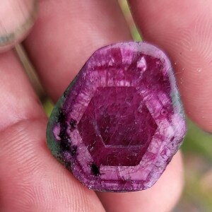 May include: A close-up of a gemstone with a deep magenta hue and a hexagonal pattern in the centre. The stone has a rough, natural texture with hints of green around the edges. The gem is held between fingers.