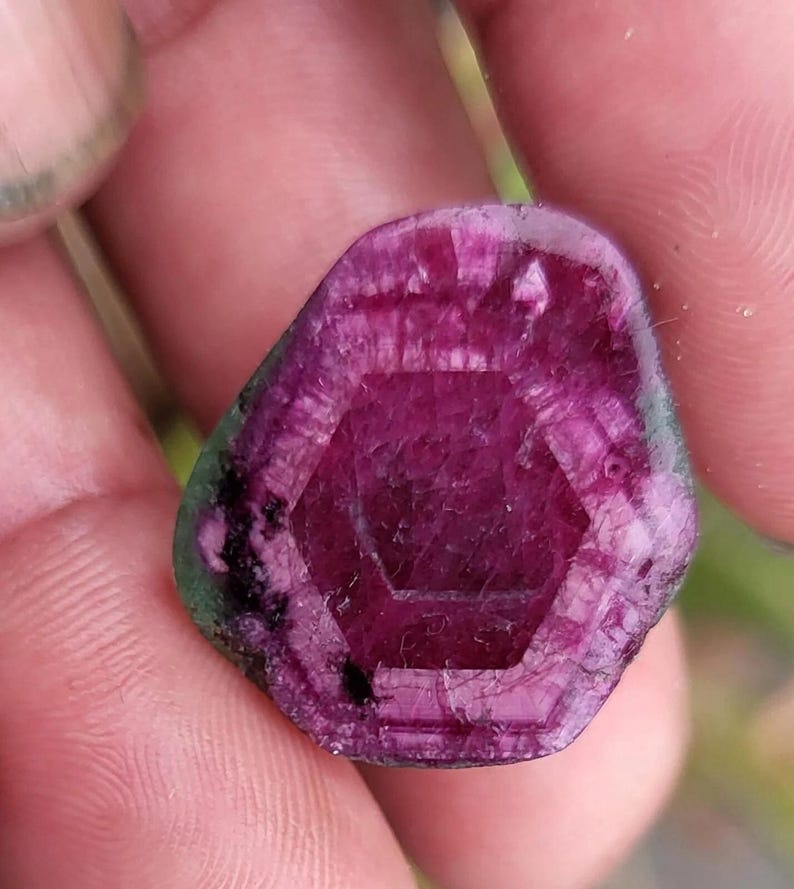 May include: A close-up of a gemstone with a deep magenta hue and a hexagonal pattern in the centre. The stone has a rough, natural texture with hints of green around the edges. The gem is held between fingers.