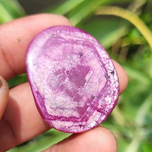 May include: A polished, oval gemstone in shades of pink and magenta. The stone features a unique pattern of internal lines and a hexagonal shape in the centre. The gemstone is held in a hand against a blurred green background.