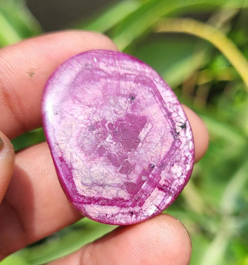 May include: A polished, oval gemstone in shades of pink and magenta. The stone features a unique pattern of internal lines and a hexagonal shape in the centre. The gemstone is held in a hand against a blurred green background.