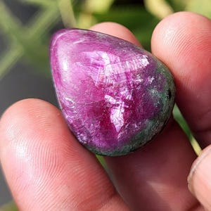 Può includere: Una gemma lucidata a forma di goccia, di colore magenta intenso, che presenta strutture cristalline interne. La pietra ha aree verdi ed è tenuta tra le dita, mostrando la sua bellezza naturale.