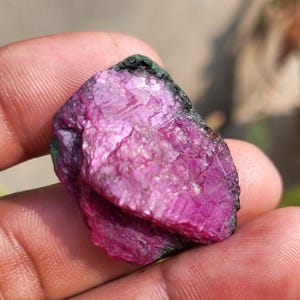 May include: A rough-cut ruby gemstone with a deep magenta hue. The surface of the stone has a textured appearance with visible crystalline structures. The edges of the ruby are dark green and black, contrasting with the vibrant pinkish-red core.