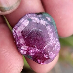 May include: A close-up of a faceted, raw gemstone with a deep magenta centre and a translucent, multi-toned edge of green and purple. The stone has a geometric, hexagonal shape in the centre and a rough, natural texture.