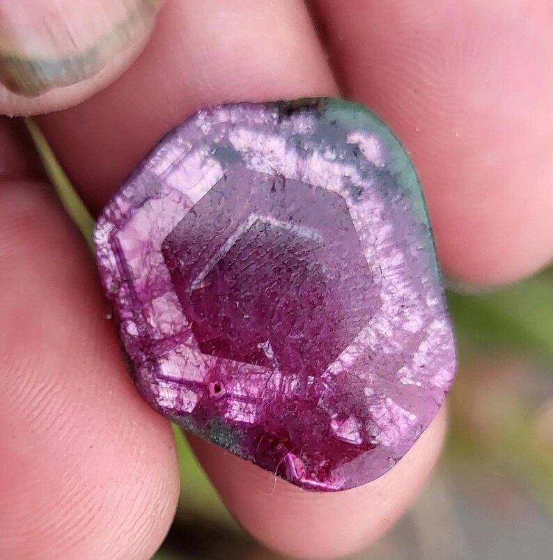 May include: A close-up of a faceted, raw gemstone with a deep magenta centre and a translucent, multi-toned edge of green and purple. The stone has a geometric, hexagonal shape in the centre and a rough, natural texture.