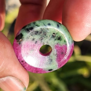 Può includere: Una perla di pietra preziosa rotonda e lucidata con un foro centrale. La perla presenta una miscela di colori, tra cui rosa intenso, verde e nero. La superficie lucida mette in risalto i motivi naturali e le variazioni della pietra.