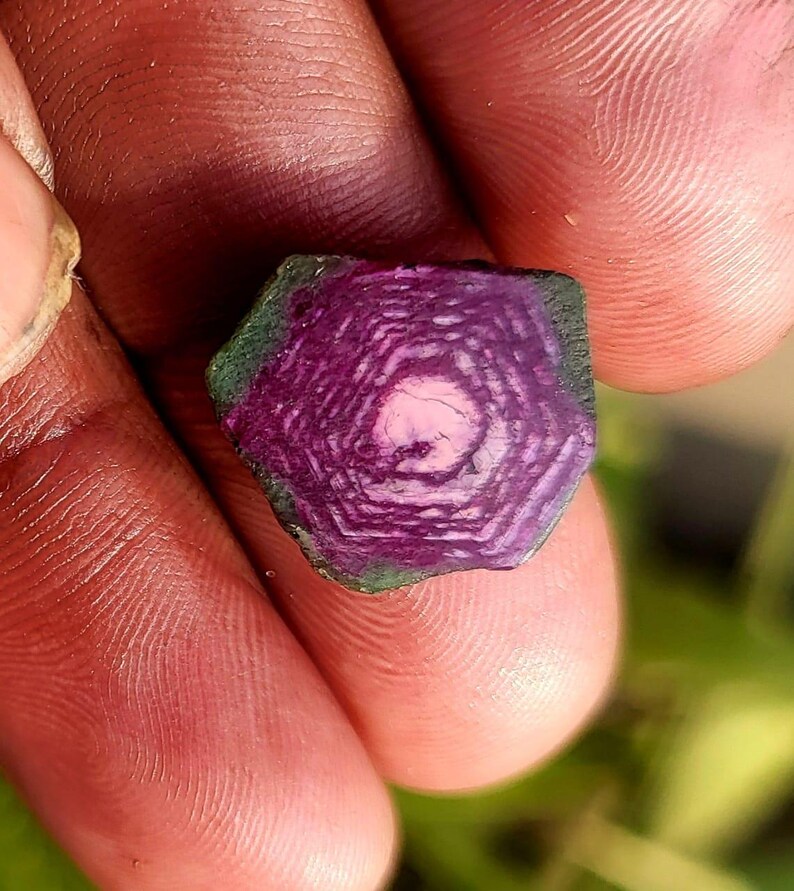 Pode incluir: Close-up de uma gema hexagonal. A pedra apresenta an&eacute;is conc&ecirc;ntricos em tons de roxo escuro e rosa, com bordas verdes. A gema &eacute; segurada entre os dedos, mostrando sua intrincada estrutura interna.