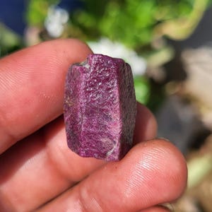 Op de afbeelding: Een ruw, zeshoekig robijnkristal met een diepe magenta-rode kleur en een getextureerd, kristallijn oppervlak. De edelsteen vertoont natuurlijke facetten en een licht onregelmatige, langwerpige vorm met zichtbare minerale insluitingen.