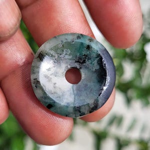 May include: A circular stone pendant with a polished surface, featuring a mottled pattern of deep forest green, teal, and black inclusions. The piece has a smooth, flat profile with a small, centered circular hole for threading or suspension.