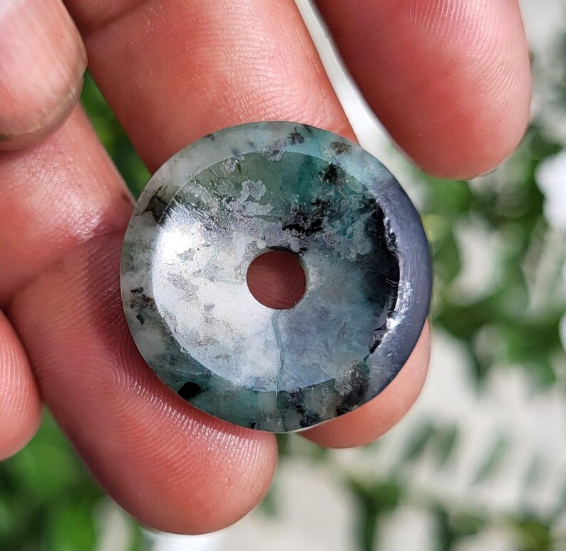 May include: A circular stone pendant with a polished surface, featuring a mottled pattern of deep forest green, teal, and black inclusions. The piece has a smooth, flat profile with a small, centered circular hole for threading or suspension.