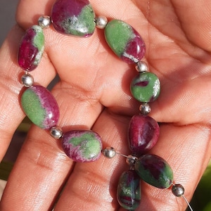 May include: A strand of gemstone beads featuring a mix of colors. The beads are a combination of deep red and green, with silver-colored spacer beads. The gemstones have an oval shape and are strung together on a thin, clear cord.