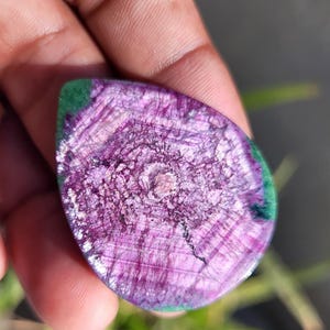 May include: A teardrop-shaped gemstone with a polished surface. The stone displays a striking purple colour with detailed radial patterns and a green border. The gemstone is held in a hand, highlighting its natural beauty and unique design.