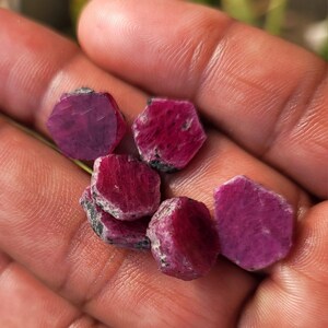 May include: A collection of raw, hexagonal-shaped ruby gemstones. The rubies display a deep red hue with some areas of darker coloration and black inclusions. The gemstones are held in a hand, showcasing their natural, unpolished state.