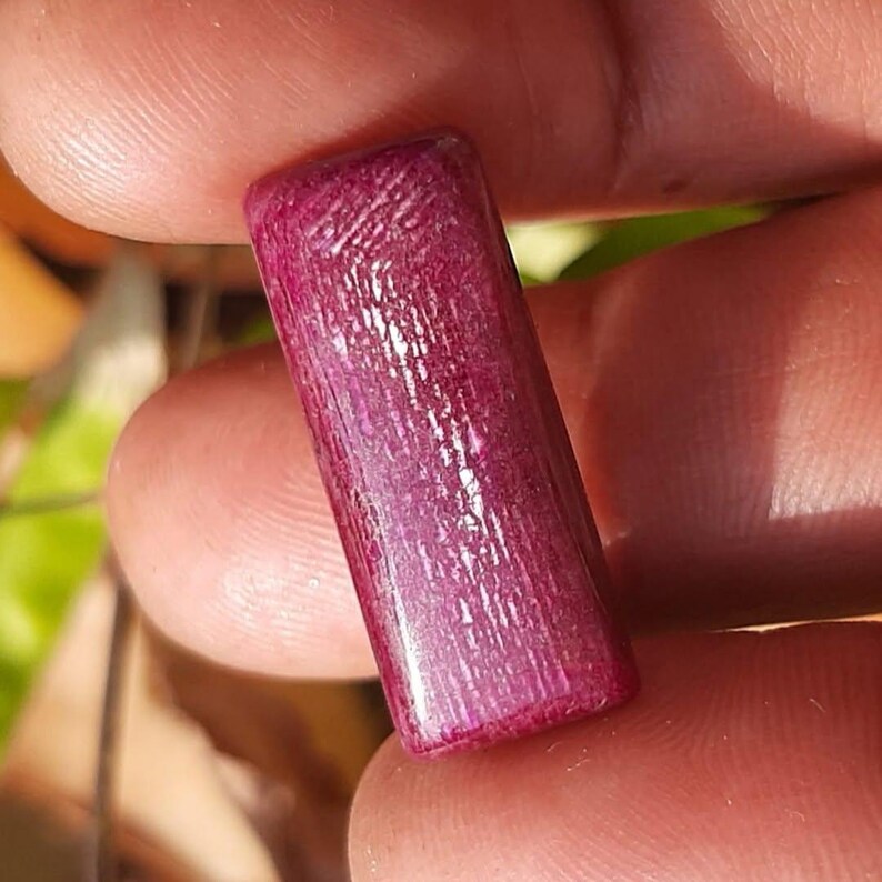 May include: A rectangular, polished gemstone in a deep magenta hue. The surface displays a textured, linear pattern, with subtle variations in color and light reflection. The gemstone is held between fingers, showcasing its size and shape.
