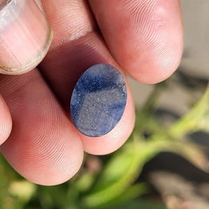 May include: Oval-shaped, blue gemstone with a speckled appearance. The stone is held between fingers, with a blurred green background. The gem's color ranges from deep blue to lighter shades, with visible inclusions.